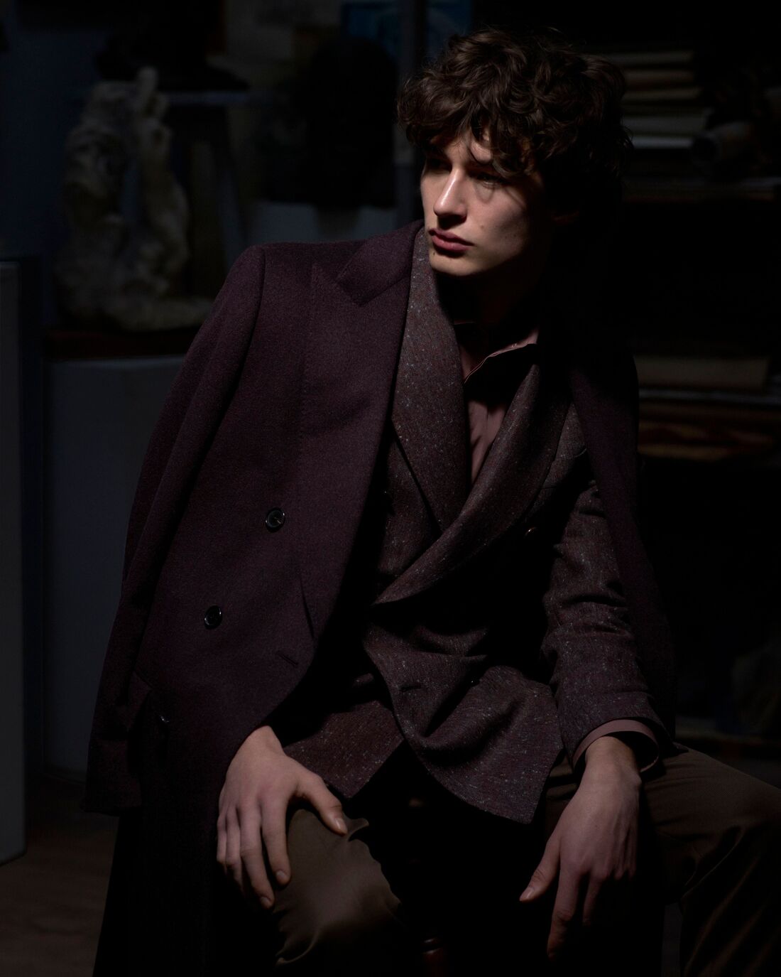 A model poses casually slightly away from the camera in a dimly lit room with a soft spotlight on their face, wearing a deep burgundy trenchcoat over their suit